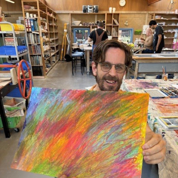 Artist Luke Kinchington proudly smiling whilst showcasing his artwork that he had just finished. It is multicoloured and vibrant. It showcases his talent for blending colours to create interesting pieces.